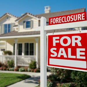 A suburban house with a 'For Sale' sign in the front yard, prominently displaying 'Foreclosure' at the top of the sign. - Nager Law Group